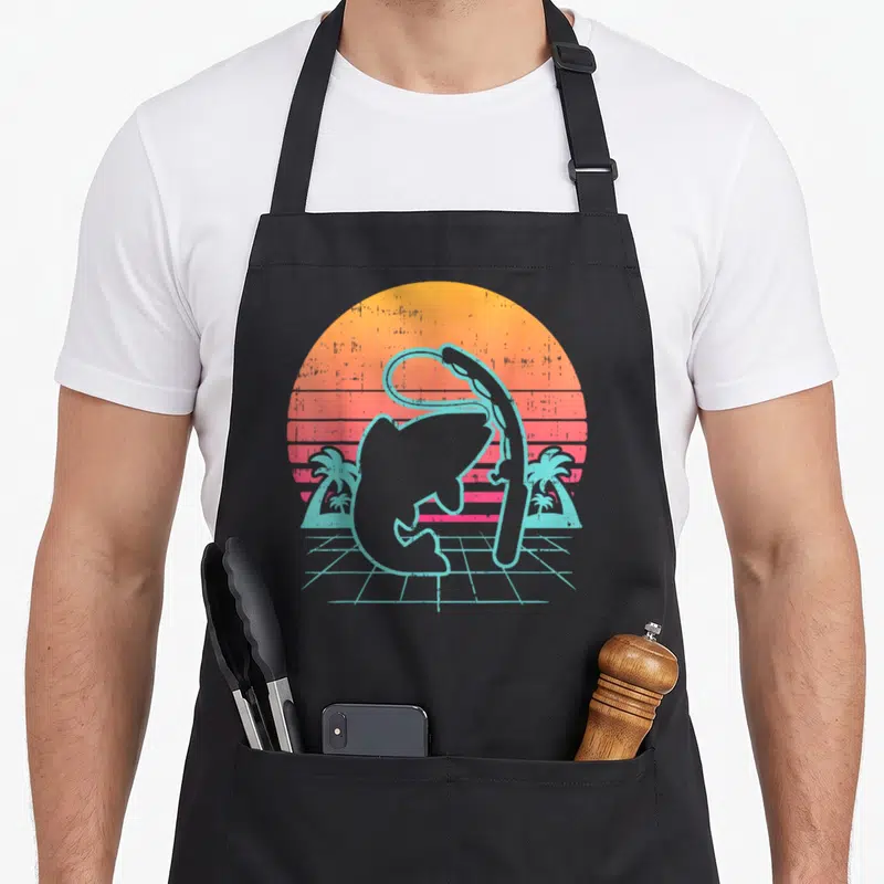 Tablier de cuisine noir avec illustration d'un poisson et d'une canne à pêche sur fond de soleil orange et palmiers turquoise.