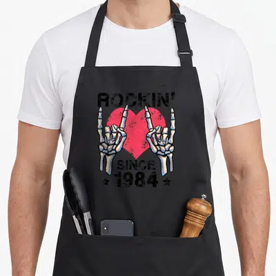 Tablier de cuisine noir avec deux mains squelettiques formant le signe du metal et un cœur rose derrière le texte Rockin Since 1984.