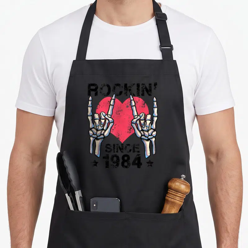 Tablier de cuisine noir avec deux mains squelettiques formant le signe du metal et un cœur rose derrière le texte Rockin Since 1984.