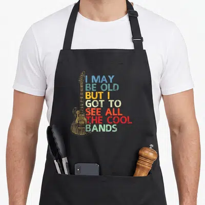 Tablier de cuisine noir avec une guitare dessinée et un texte multicolore sur fond gris uni.
