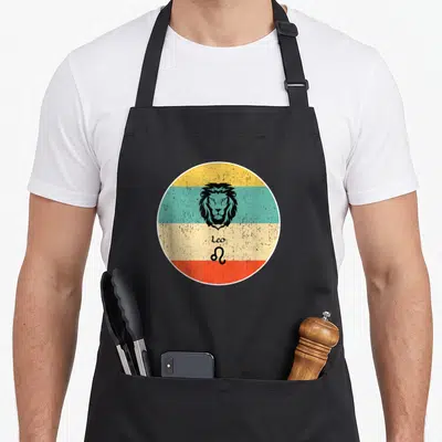 Tablier de cuisine noir avec un motif circulaire rétro représentant la tête d'un lion et le signe astrologique Leo.