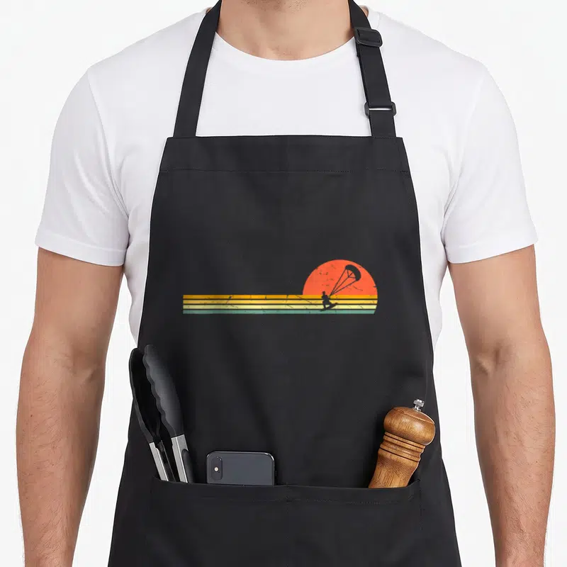 Tablier de cuisine noir avec illustration rétro de kitesurfeur sur fond de soleil orange et bandes horizontales colorées.