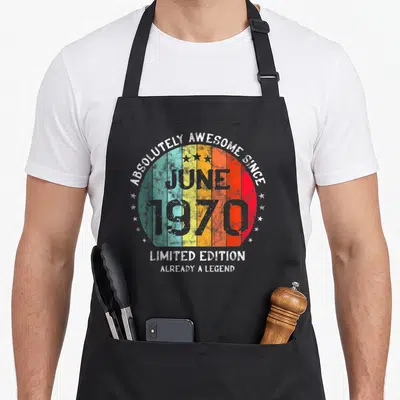 Tablier de cuisine noir avec motif circulaire rétro multicolore, étoiles blanches et texte gris mentionnant juin 1970.