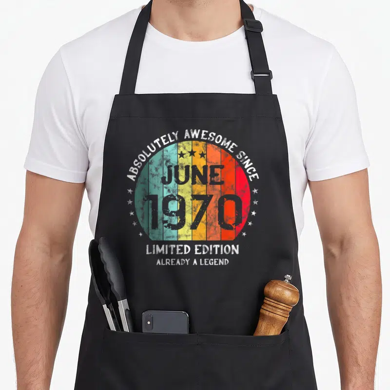 Tablier de cuisine noir avec motif circulaire rétro multicolore, étoiles blanches et texte gris mentionnant juin 1970.