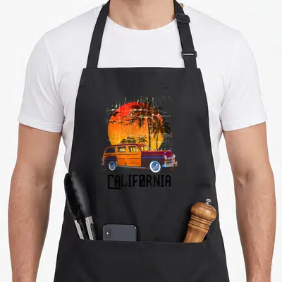 Tablier de cuisine noir avec illustration d'une voiture Woodie rouge et bois, soleils orange et palmiers au fond.