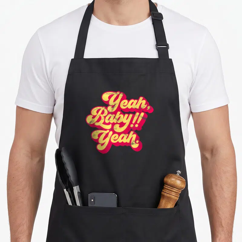 Tablier de cuisine noir avec inscription Yeah Baby Yeah en lettres jaunes et roses style rétro.