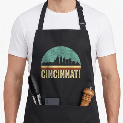 Tablier de cuisine noir avec motif circulaire turquoise, bandes orange et texte CINCINNATI en lettres jaunes texturées.