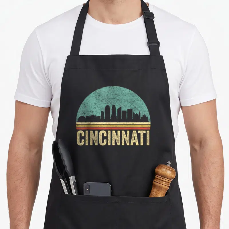Tablier de cuisine noir avec motif circulaire turquoise, bandes orange et texte CINCINNATI en lettres jaunes texturées.
