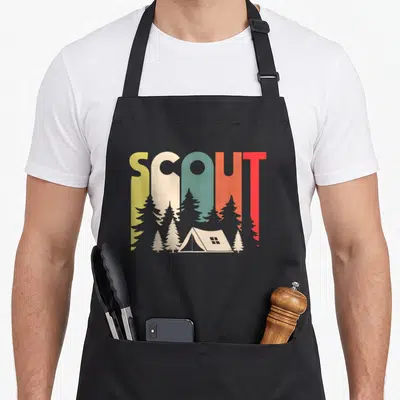 Tablier de cuisine noir avec l'inscription Scout en lettres multicolores au-dessus d'une tente et de sapins stylisés.