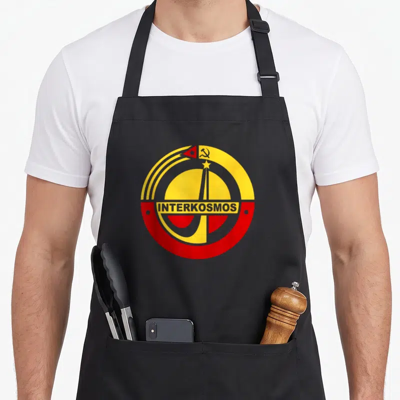Tablier de cuisine noir avec le logo circulaire rouge et jaune Interkosmos représentant un lanceur spatial soviétique.