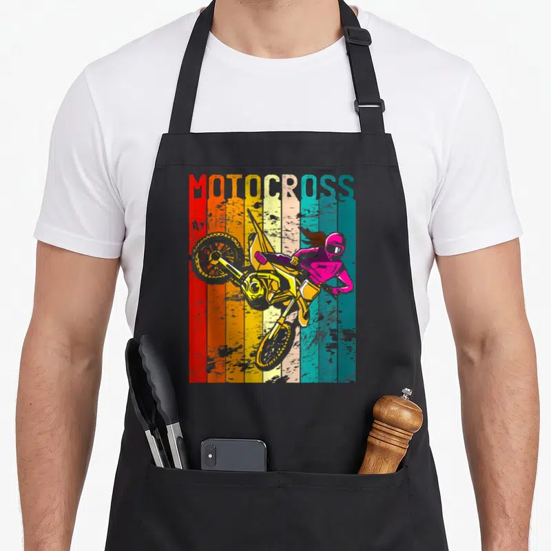 Tablier de cuisine noir avec illustration rétro d'une pilote de motocross sur moto jaune et casque rose devant des bandes de couleur.