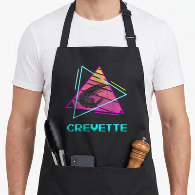 Tablier de cuisine noir avec une crevette rose inscrite dans des triangles cyan et magenta sous le mot CREVETTE en lettres pixelisées.