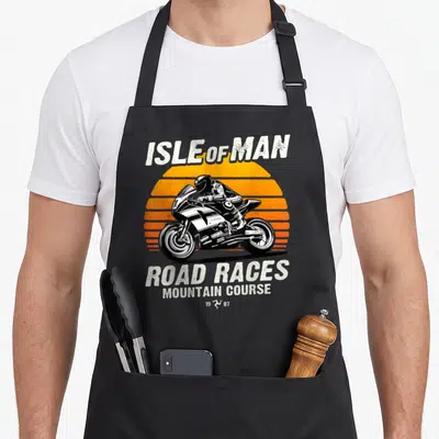 Tablier de cuisine noir avec illustration rétro d'une course de moto Isle of Man sur fond de soleil orange strié.