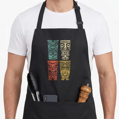 Tablier de cuisine noir avec quatre masques Tiki polynésiens colorés en turquoise, beige, rouge et jaune disposés en grille.
