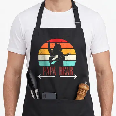 Tablier de cuisine noir avec illustration d'un ours jouant de la guitare devant un soleil coloré et l'inscription Papa Bear en rouge.