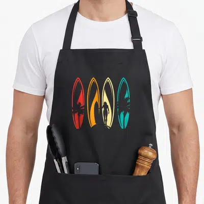 Tablier de cuisine noir avec quatre planches de surf colorées et silhouettes de surfeur et palmiers en style rétro.