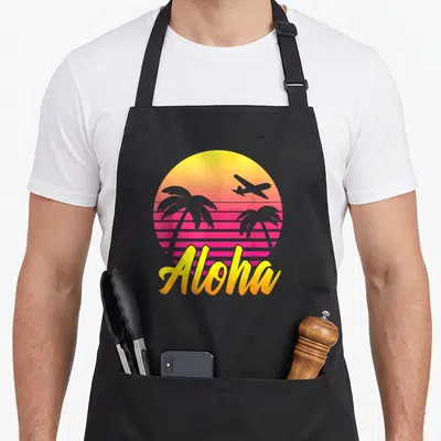 Tablier de cuisine noir avec illustration d'un soleil couchant rose et jaune, palmiers, avion et le mot Aloha en écriture cursive dorée.