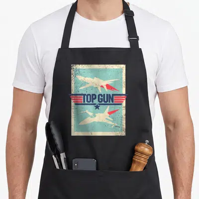 Tablier de cuisine noir avec illustration rétro Top Gun montrant deux jets et un logo aux ailes bleues et rouges.