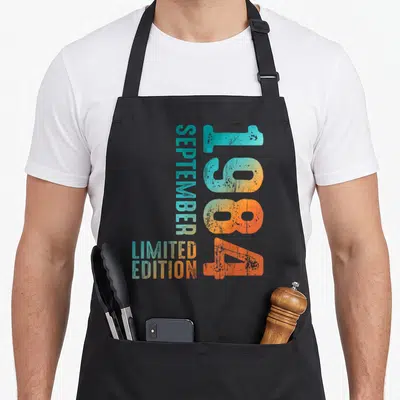 Tablier de cuisine noir avec l'inscription Septembre 1984 en typographie rétro dégradée orange et turquoise.