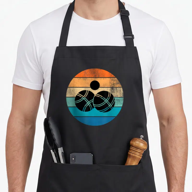 Tablier de cuisine noir avec illustration circulaire rétro de deux boules de pétanque noires sur fond de bandes colorées orange et bleues.