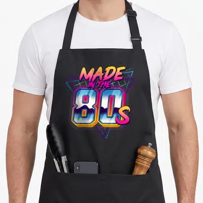 Tablier de cuisine noir avec l'inscription « Made in the 80s » en lettres colorées et motifs géométriques bleus et roses.