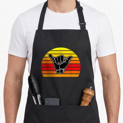Tablier de cuisine noir avec un signe Shaka noir sur un soleil rayé orange et jaune au design rétro.