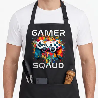Tablier de cuisine noir avec une illustration de manette de jeu vidéo multicolore et le texte Gamer Squad en blanc.