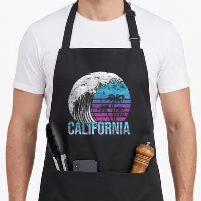 Tablier de cuisine noir avec illustration circulaire de vague et palmiers au-dessus du mot California en lettres bleues striées.