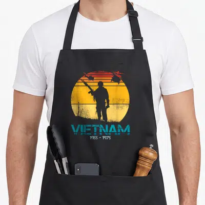 Tablier de cuisine noir avec silhouette de soldat, hélicoptères et texte Vietnam 1955-1975 sur fond de soleil orange strié.