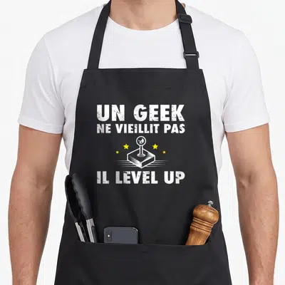 Tablier de cuisine noir avec un joystick blanc, des étoiles jaunes et le texte Un Geek Ne Vieillit Pas Il Level Up.