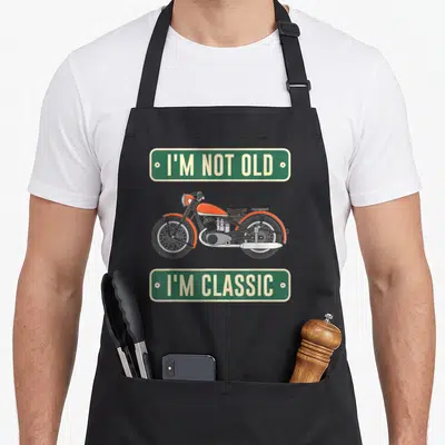 Tablier de cuisine noir avec illustration de moto orange et panneaux verts portant des textes en anglais.
