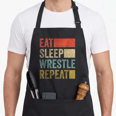 Tablier de cuisine noir avec inscription Eat Sleep Wrestle Repeat en lettres colorées style rétro 70s.
