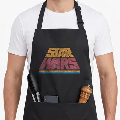 Tablier de cuisine noir avec logo Star Wars Classic Retro Slant Logo en orange, rose et bleu sur fond uni.