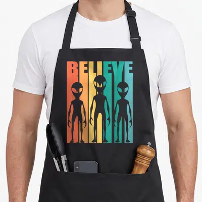 Tablier de cuisine noir avec trois aliens gris devant le mot Believe en bandes colorées orange jaune et vert turquoise