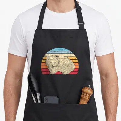 Tablier de cuisine noir avec illustration d'un wombat devant un soleil rayé aux couleurs pastel bleues, oranges et rouges.