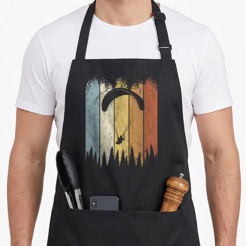 Tablier de cuisine noir avec silhouette de parapente sur fond de bandes colorées et silhouettes d'arbres en bas.