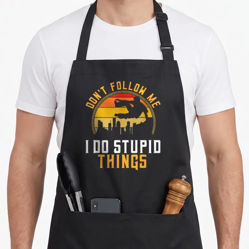 Tablier de cuisine noir avec inscription Ne me suis pas je fais des choses stupides et silhouette de saut urbain sur fond orange.