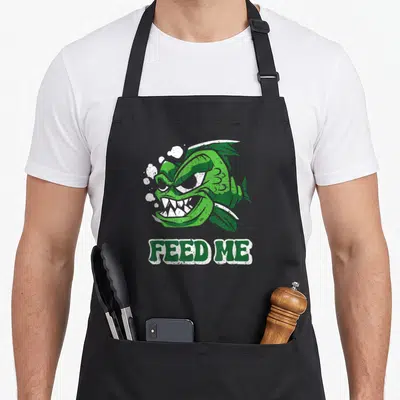 Tablier de cuisine noir avec illustration d'un piranha vert rétro et le texte FEED ME en lettres capitales blanches et vertes.