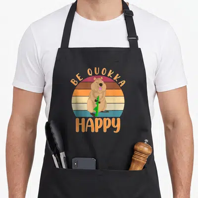 Tablier de cuisine noir avec illustration rétro d'un quokka souriant et le texte Be Quokka Happy.