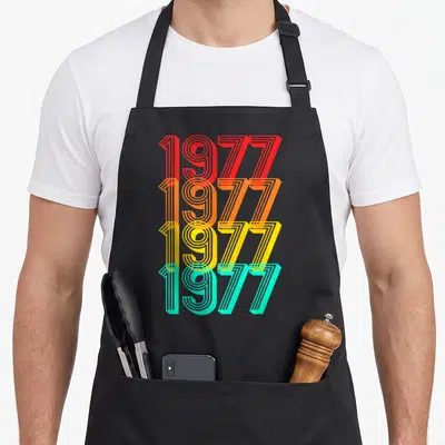 Tablier de cuisine noir avec quatre lignes de chiffres 1977 aux couleurs dégradées orange jaune et turquoise.