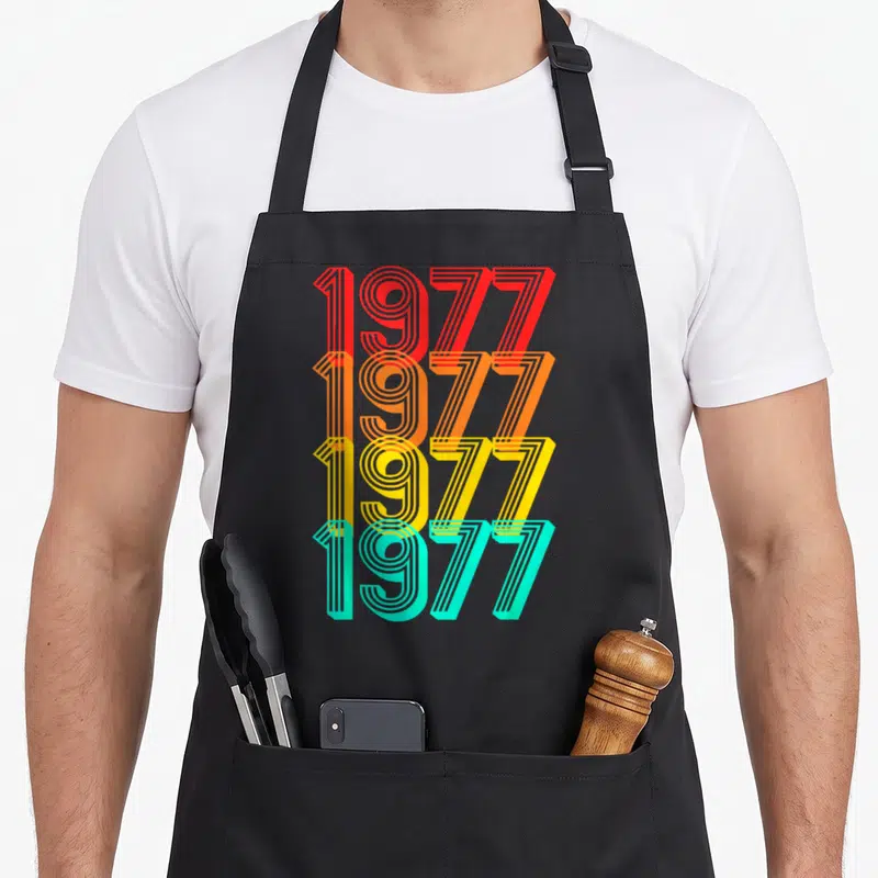 Tablier de cuisine noir avec quatre lignes de chiffres 1977 aux couleurs dégradées orange jaune et turquoise.