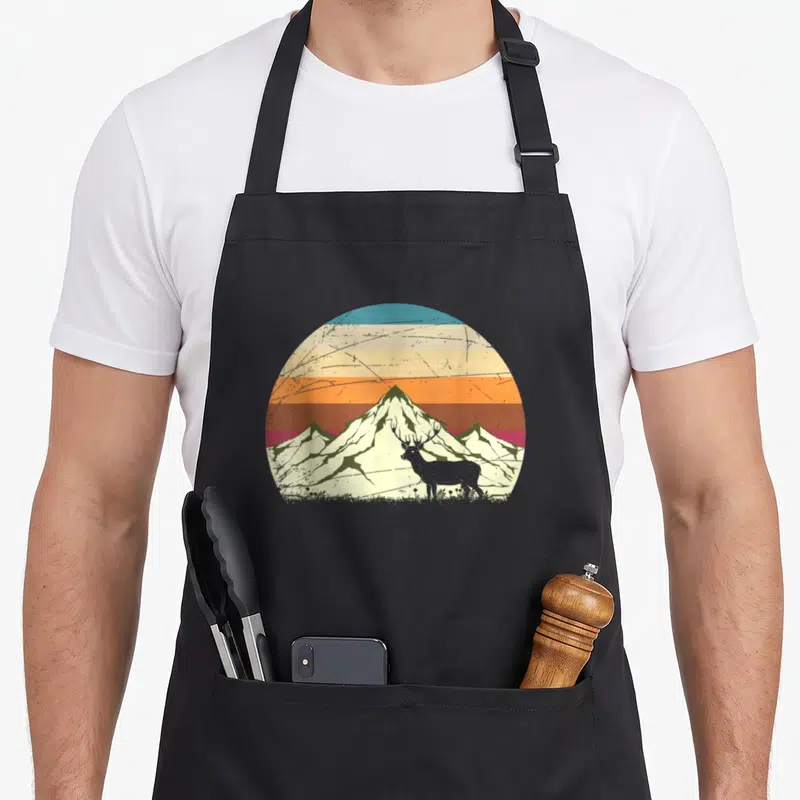 Tablier de cuisine noir avec illustration circulaire retro montrant un cerf devant des montagnes aux couleurs chaudes.