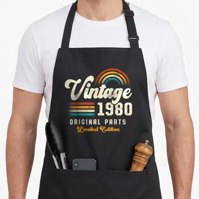 Tablier de cuisine noir avec inscription Vintage 1980 Original Parts Limited Edition et arc-en-ciel coloré sur le devant.