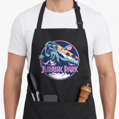 Tablier de cuisine noir avec illustration circulaire rétro Jurassic Park montrant un T-Rex turquoise et des palmiers sous un ciel coloré.