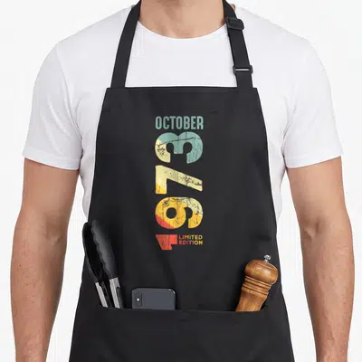Tablier de cuisine noir avec le texte vertical Octobre 1973 Limited Edition dans des tons pastel délavés.