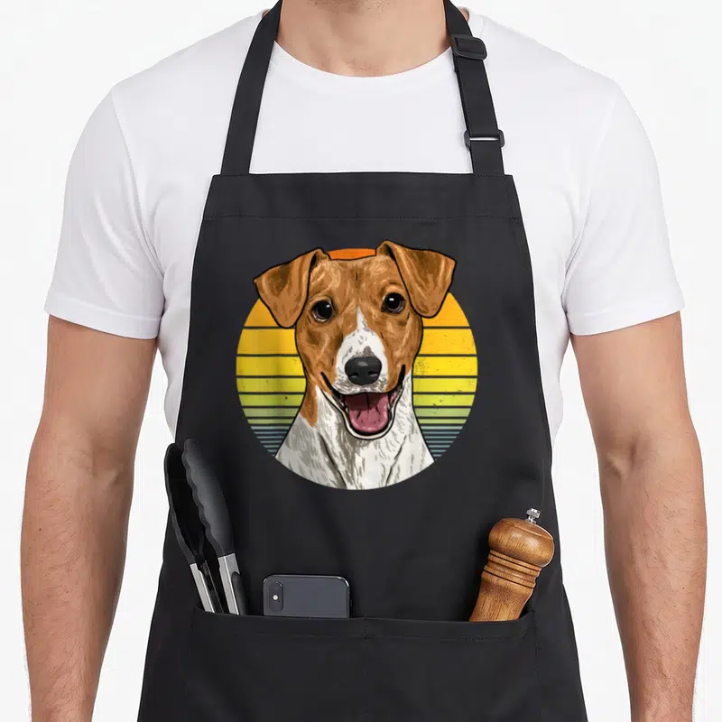 Tablier de cuisine noir avec illustration circulaire d'un Jack Russel Terrier sur fond de coucher de soleil rétro jaune et bleu.