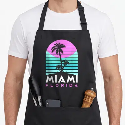 Tablier de cuisine noir avec illustration MIAMI FLORIDA, palmiers et soleil rose néon sur fond de lignes turquoise horizontales.