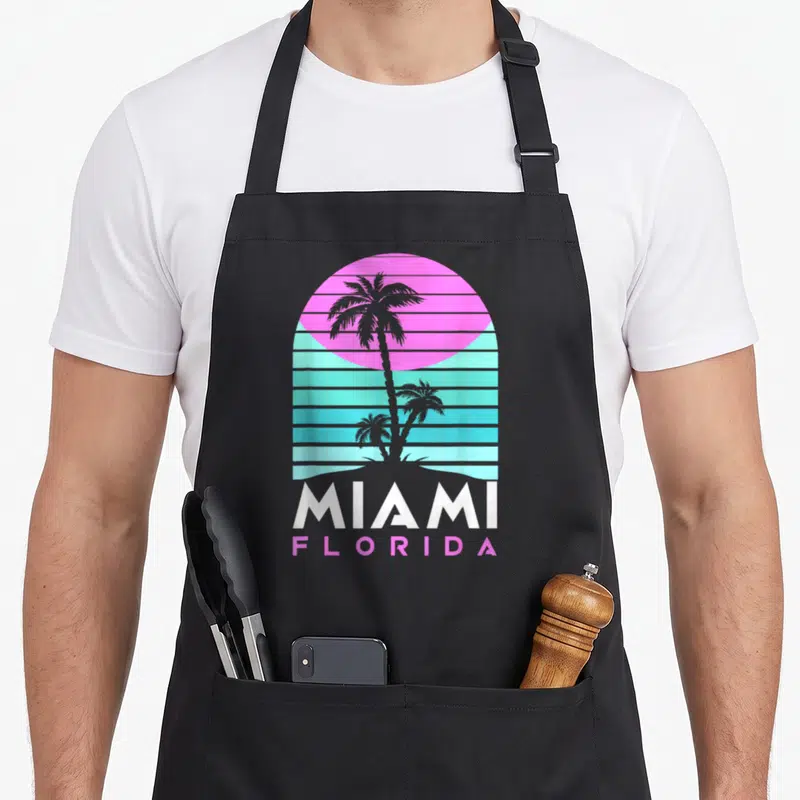 Tablier de cuisine noir avec illustration MIAMI FLORIDA, palmiers et soleil rose néon sur fond de lignes turquoise horizontales.