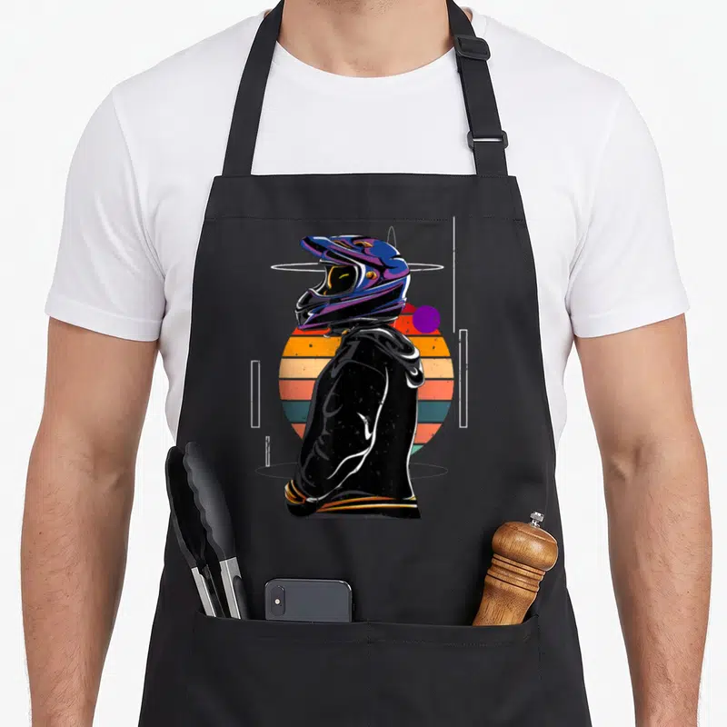 Tablier de cuisine noir avec illustration d'un casque de moto coloré devant un soleil couchant rayé orange et bleu.