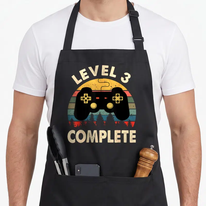 Tablier de cuisine noir avec manette de jeu, soleil rayé et texte Level 3 Complete en style rétro.